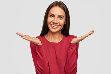 Positive hesitant cheerful young European tanned woman with long dark hair, raises palms in bewilderment, cant make decision between something pleasant, isolated over white background. So what?