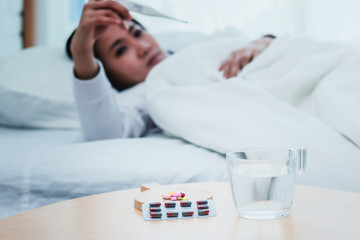 Closeup on pills and glass of water on table and Asian women sleep sick on bed in bed room.color tone