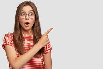 Its incredible, look! Astonished dark haired young woman being amazed by what is near her, points with index finger aside, opens mouth from amazement, wears round glasses and striped t shirt.