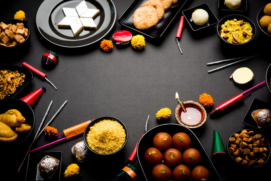 Diwali sweets and snacks arranged in a group with Diya or oil lamp, flowers and Fire Crackers or Patakhe over moody background, selective focus