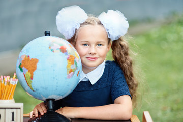 Portrait of a beautiful young first-grader sitting at a desk on the background autumn park. Farewell Bell. day of knowledge. beginning of the school year. school equipment