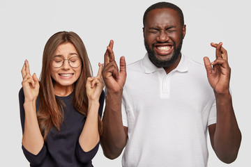 Photo of cheerful dark skinned man, positive European woman cross fingers, have charming smiles, believe in good fortune before important event, pose against white background. Diversity concept - obrazy, fototapety, plakaty