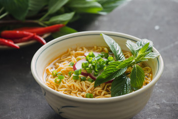 Instant noodles in bowl with fresh herbs, garnish of cilantro and Asian basil, lemon, lime on dark stone background