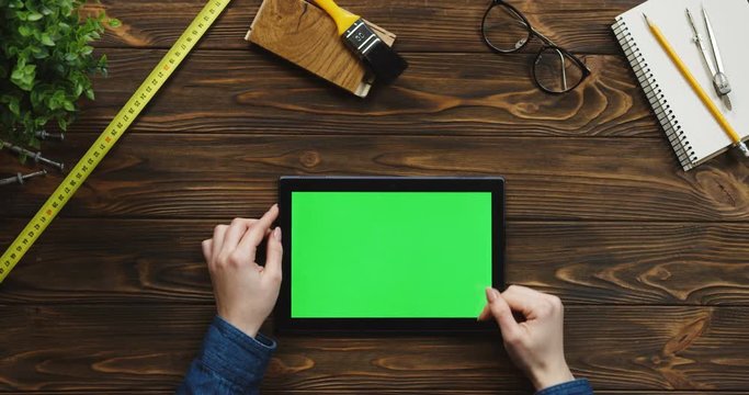 View from above on the black horizontal tablet device on the wooden desk with craftsman equipment, Caucasian female hands scrolling and tapping on the green screen. Chroma key.