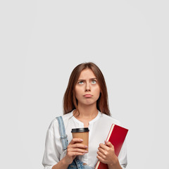 Indoor shot of displeased gloomy student holds disposable cup of coffee, notepad, looks with bewilderment upwards, frowns eyebrows, free space above for your promotional content. Learning concept