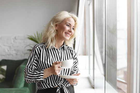 People, lifestyle, food and beverage concept. Portrait of fashionable elegant sixty year old lady wearing stylish striped blouse, halving relaxed happy look, enjoying hot drink by the window