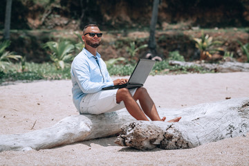 young businessman in sunglasses blue shirt and shorts working with laptop on the beach