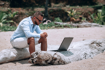young businessman in sunglasses blue shirt and shorts working with laptop on the beach