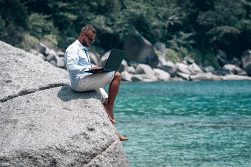 young businessman in sunglasses blue shirt and shorts working with laptop on the beach