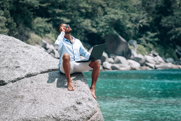 young businessman in sunglasses blue shirt and shorts working with laptop on the beach