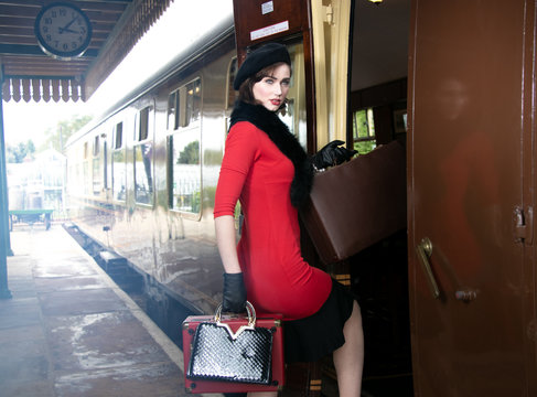 vintage attractive female wearing red dress and black beret with suitcases on platform of train station