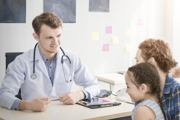 Pediatrician (doctor) man examining child with stethoscope at surgery .Mother Caucasian and kid smiling in hospital room.Copy space.