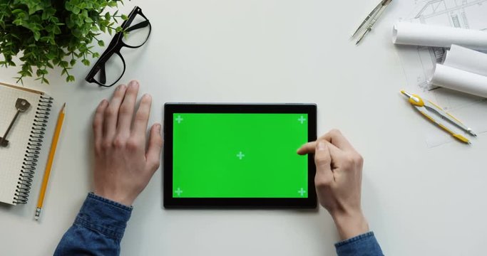 Top view on the male hands scrolling and tapping on the green screen of the black horizontal tablet computer on the designer or architect table. Chroma key. Tracking motion.