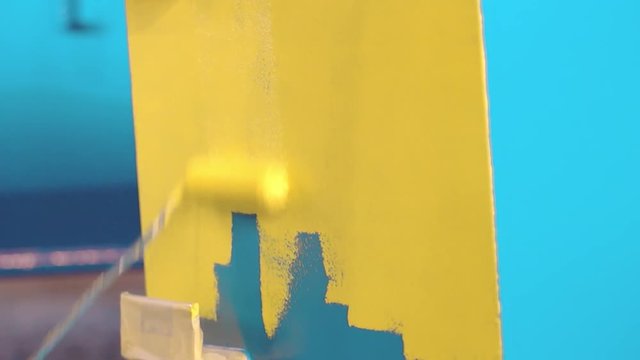 Man is painting large blue metal detail by hand roller to yellow color. Painter at the factory. Professional workshop. Heavy industry servis and metal construction factory
