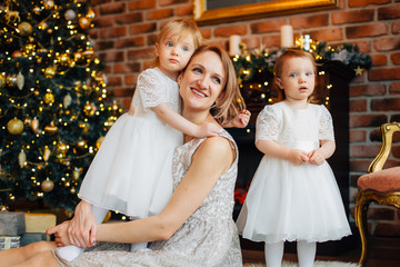 Mother with two cute little daughters in white dress sitting on the floor near christmas tree. Smiling woman playing with cheerful girls sisters. Happy new year