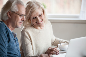 Senior happy couple enjoying using laptop eating breakfast together, smiling elderly middle aged family shopping online, reading discussing internet news, buying travel tour on computer site at home