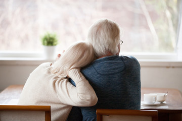 Rear view at senior grey haired loving caring family couple embracing relaxing at home together enjoying peaceful morning breakfast looking at window view thinking of future feeling calm nostalgic.