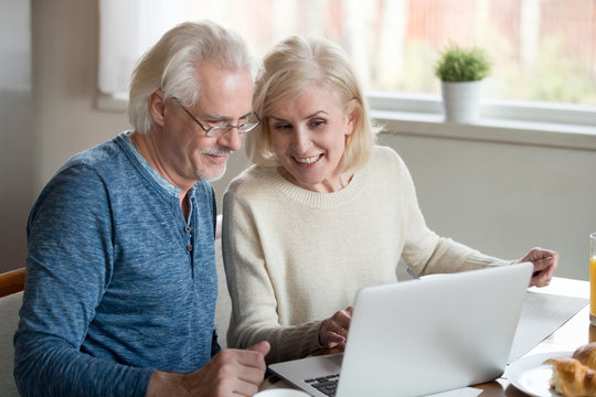 Happy old family couple talking using laptop having breakfast together, surprised excited senior woman looking at computer screen showing smiling middle aged husband online shopping sale on web site