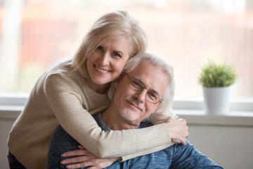 Smiling caring middle aged wife embracing senior husband at home, happy old woman hugging loving...
