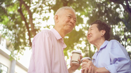Happy Asian elderly couple morning walk in green city while drinking coffee and talking