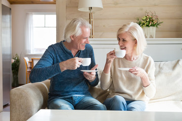 Happy senior old couple sitting on sofa in living room talking enjoying tea at home, smiling middle aged mature family chatting laughing drinking coffee having fun pleasant conversation together