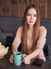 Lifestyle portrait of beautiful cute blonde woman with long hair blue eyes pastel outfit enjoying tea sitting in the cozy cafe near the window with shy smiling