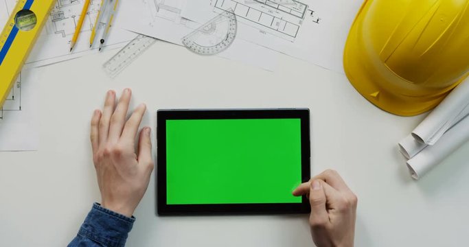 View from above on the male hands scrolling and tapping on the chroma key screen of the black horizontal tablet device on the builder desk with drawings stuff and helmet. Green screen.