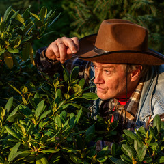 Senior adult man or a farmer, in a brown cowboy hat, looks out of the bushes and makes a grimace. Concept of active leisure for middle-aged and older people. For instagram format. Square.