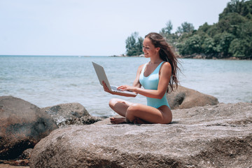 Fototapeta na wymiar Young cute woman using laptop and sitting on stone near sea, empty morning beach. Concept of modern technology and working on nature, summer vacations.