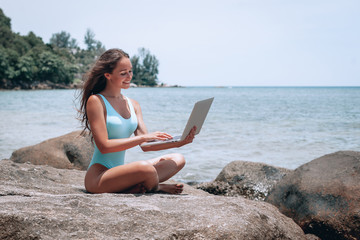 Young caucasian woman working with laptop on empty beach and sitting on stone. Concept of resting on morning sea and summer vacations, modern technology.