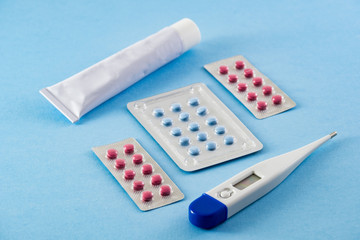Blister packs of blue and pink pills, white ointment tube  and electronic digital thermometer on blue background