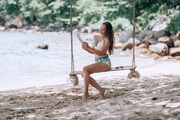 Young fit girl in denim shorts and a white bra swinging on a swing on the shore of the Andaman sea in Phuket. that the writes and reads in Notepad