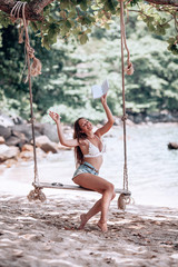 Young fit girl in denim shorts and a white bra swinging on a swing on the shore of the Andaman sea in Phuket.With his hand raised up. Happy