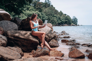 Young happy woman using laptop and sitting on stone near sea, empty morning beach. Concept of modern technology and working on nature, summer vacations.