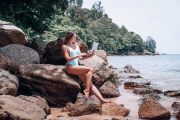 Young american woman in blue swimsuit working with laptop on empty beach and sitting on stone. Concept of resting on morning sea and summer vacations, modern technology.