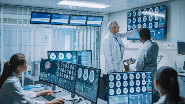 Team of Professional Medical Scientists Work in the Brain Research Laboratory. Neurologists / Neuroscientists Surrounded by Monitors Showing CT, MRI Scans Having Discussion and Working on PС