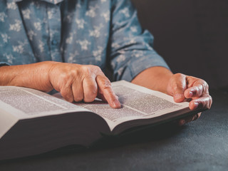 Old woman reading thick book at home. Grandmother with Bible. Concentrated elderly pensioner with wrinkles on hands attentively follows finger on paper page in library