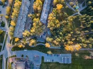 Aerial view of strung five-story buildings in the city center among tall trees with green and yellow leaves with cars on a parking on a sunny autumn day at sunset
