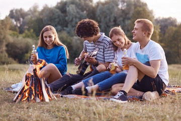 Fototapeta premium Joyful teenagers sit closely to each other on ground near fire, have picnic together, play acoustic guitar, celebrate something, have outdoor party, drink cold energetic drinks. Leisure concept