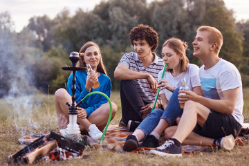 Happy smiling company have picnic together, smoke hookah, sits near bonfire, communicate with each...