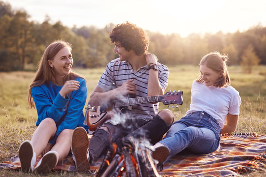 Two cheerful cute girls spend free time with handsome curly male who plays guitar and entertains them, pose together on plaid near campfire in field durring sunny weather. Companionship concept