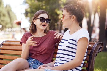 Two girl have friendly conversation in park, discuss something actively, wear trendy sunglasses, pose on wooden bench, enjoy sunny day, have walk outdoor. People, friendship, communication concept