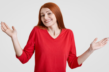Pretty redhaired woman with cheerful expression, shruggs shoulders with hesitation, has doubtful...
