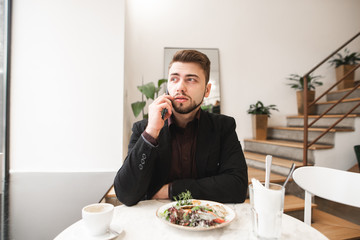 Business man sitting at a restaurant with salad and a cup of coffee on the table, talking on the smartphone and looking away.Man in a suit and a beard calls the phone in a cozy restaurant.