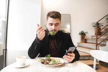 Fototapeta na wymiar Business man use a smartphone during breakfast in the restaurant. Attractive business man sits in a cozy restaurant, eating salad greens and using a smartphone.