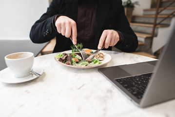 Man in a suit eats a salad with a fork and a knife, a cup of coffee and a laptop is on the table in the restaurant. A busy man dishes in a cafe and uses a notebook. Copyspace