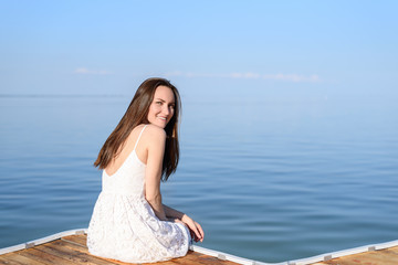 Fototapeta na wymiar Brunette sitting on the pier in a white sundress, smiling, looking at the camera in half a turn on the background of the sea, rear view, copy space