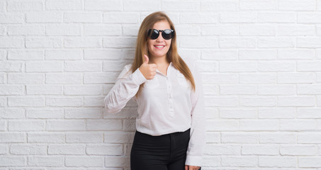 Young adult business woman standing over white brick wall wearing sunglasses happy with big smile doing ok sign, thumb up with fingers, excellent sign