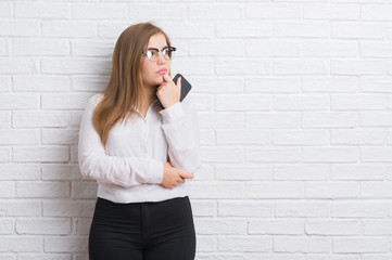 Young adult business woman over white brick wall sending message using smartphone serious face...
