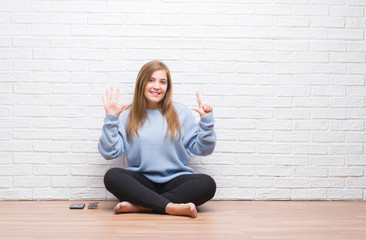 Young adult woman sitting on the floor in autumn over white brick wall showing and pointing up with fingers number seven while smiling confident and happy.